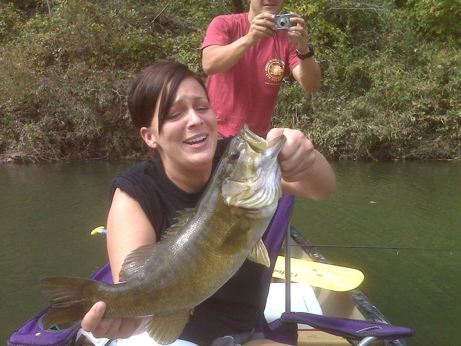 Woman holding a large fish while sitting in a small boat on a river, with another person standing behind her.