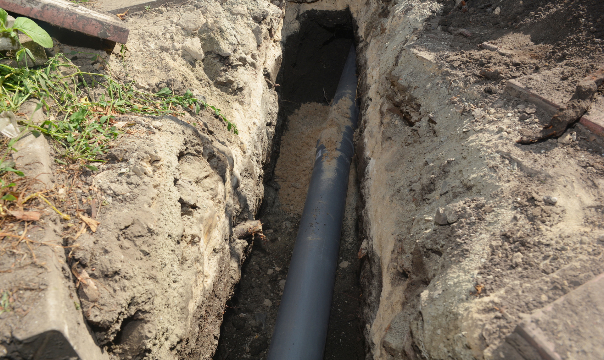 Black pipe laid in a narrow dirt trench outdoors, surrounded by excavated soil.