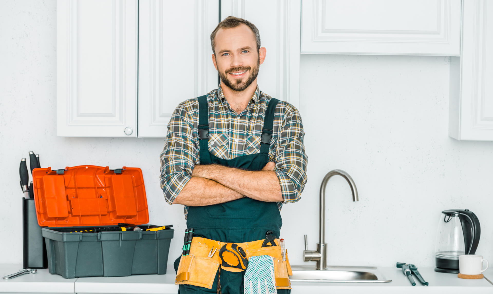 Plumber in a kitchen with tools and a toolbox, standing with arms crossed beside a sink. Plumber in a kitchen with tools and a toolbox, standing with arms crossed beside a sink.