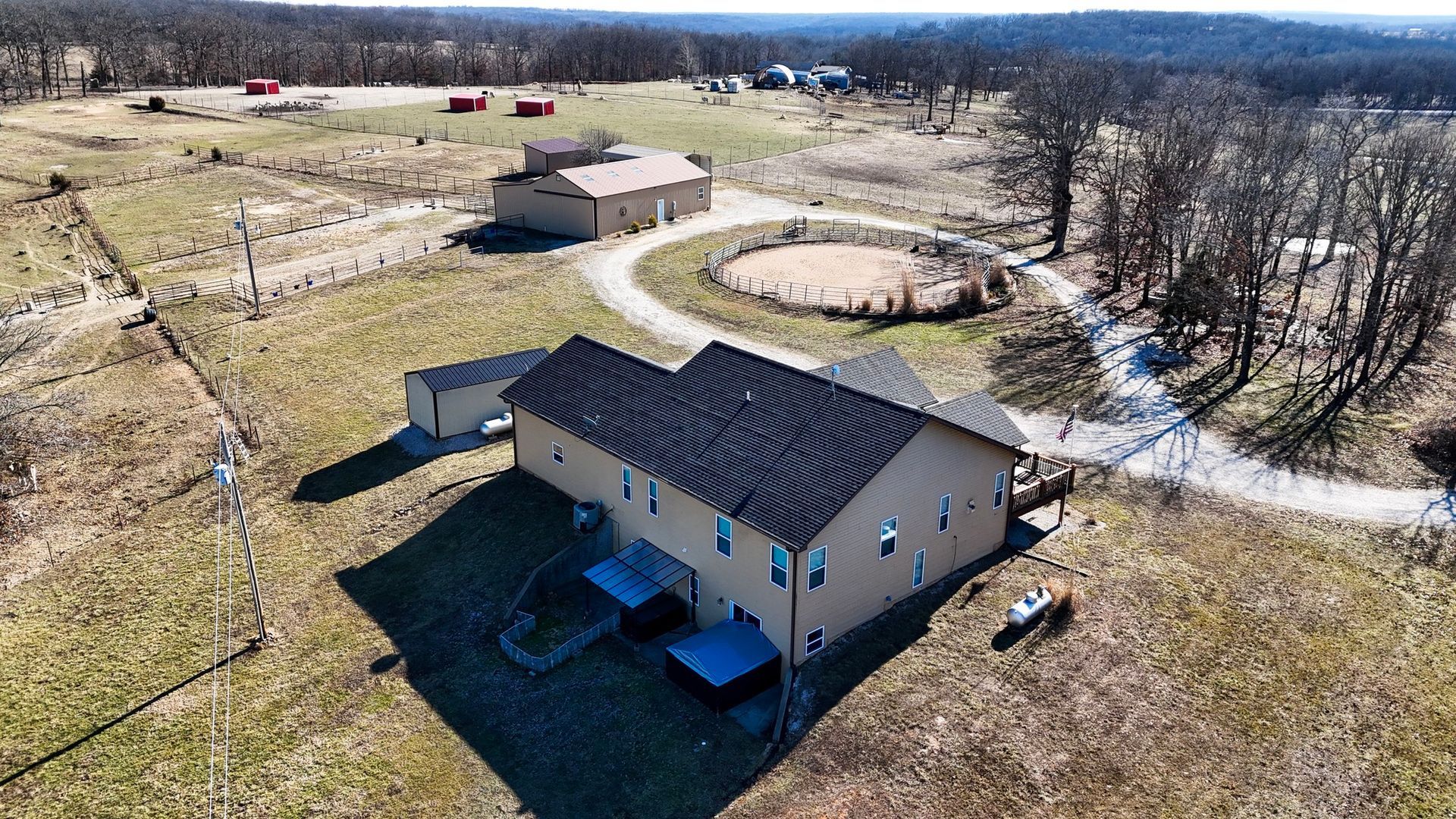 An aerial view of a light brown house and adjacent barn on a rural property with a circular riding ring and open fields.