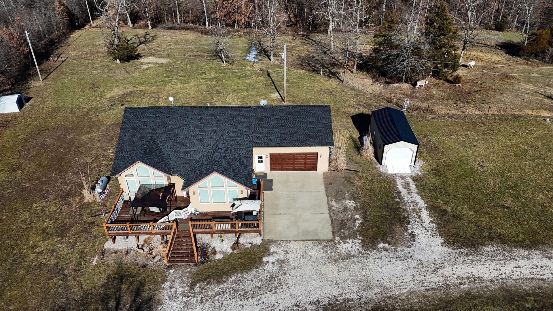 An aerial view of a single-story beige house with a dark roof, a wooden deck, a concrete driveway, and a detached shed.