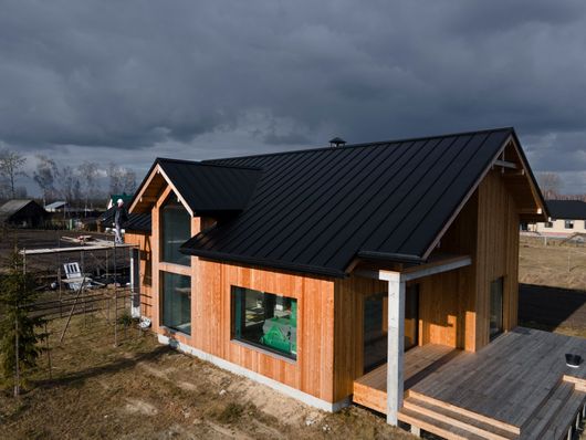 Wooden house with black roof, large windows, and a deck under a cloudy sky.