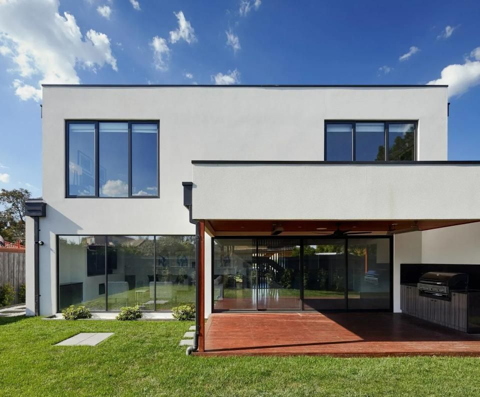 Modern two-story white house with large windows, a red patio, and a grassy lawn under a blue sky.