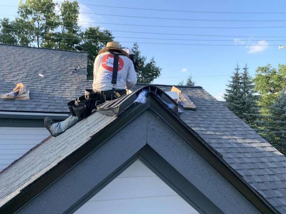 Roofer on a roof, working on dark trim and gray shingles, wearing a hard hat and tool belt.