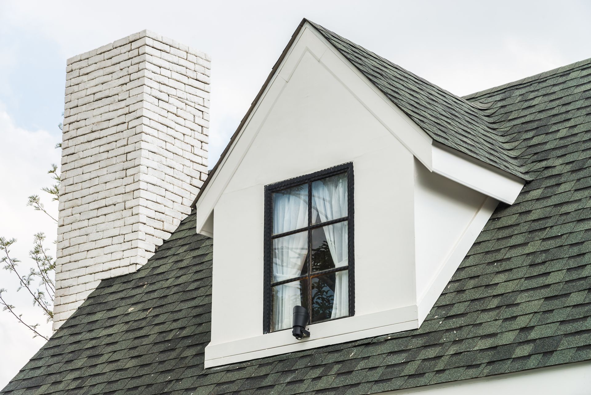 White dormer with window on a green shingled roof, a brick chimney is in the background.