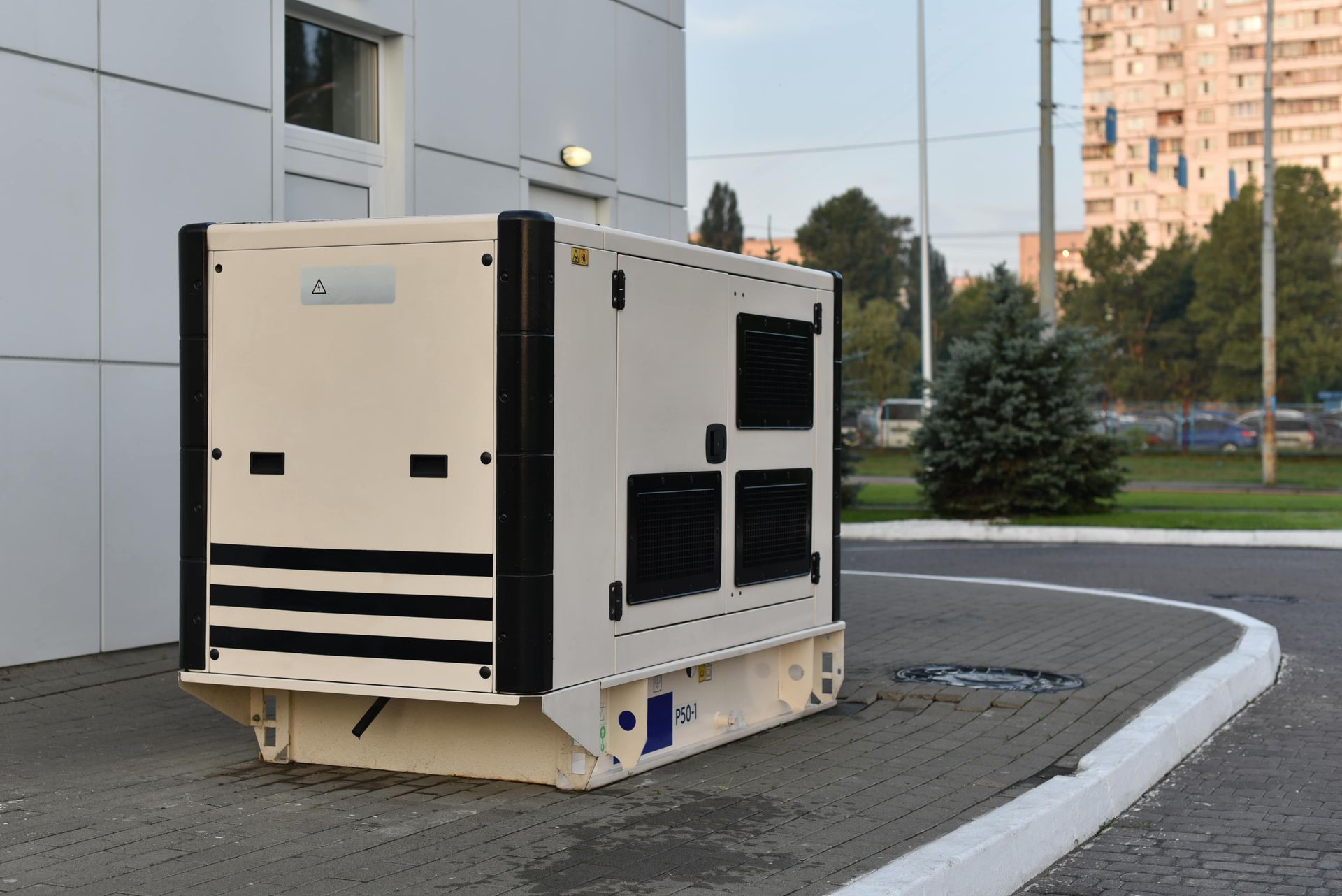 A beige generator with black accents sits on a paved area beside a building.