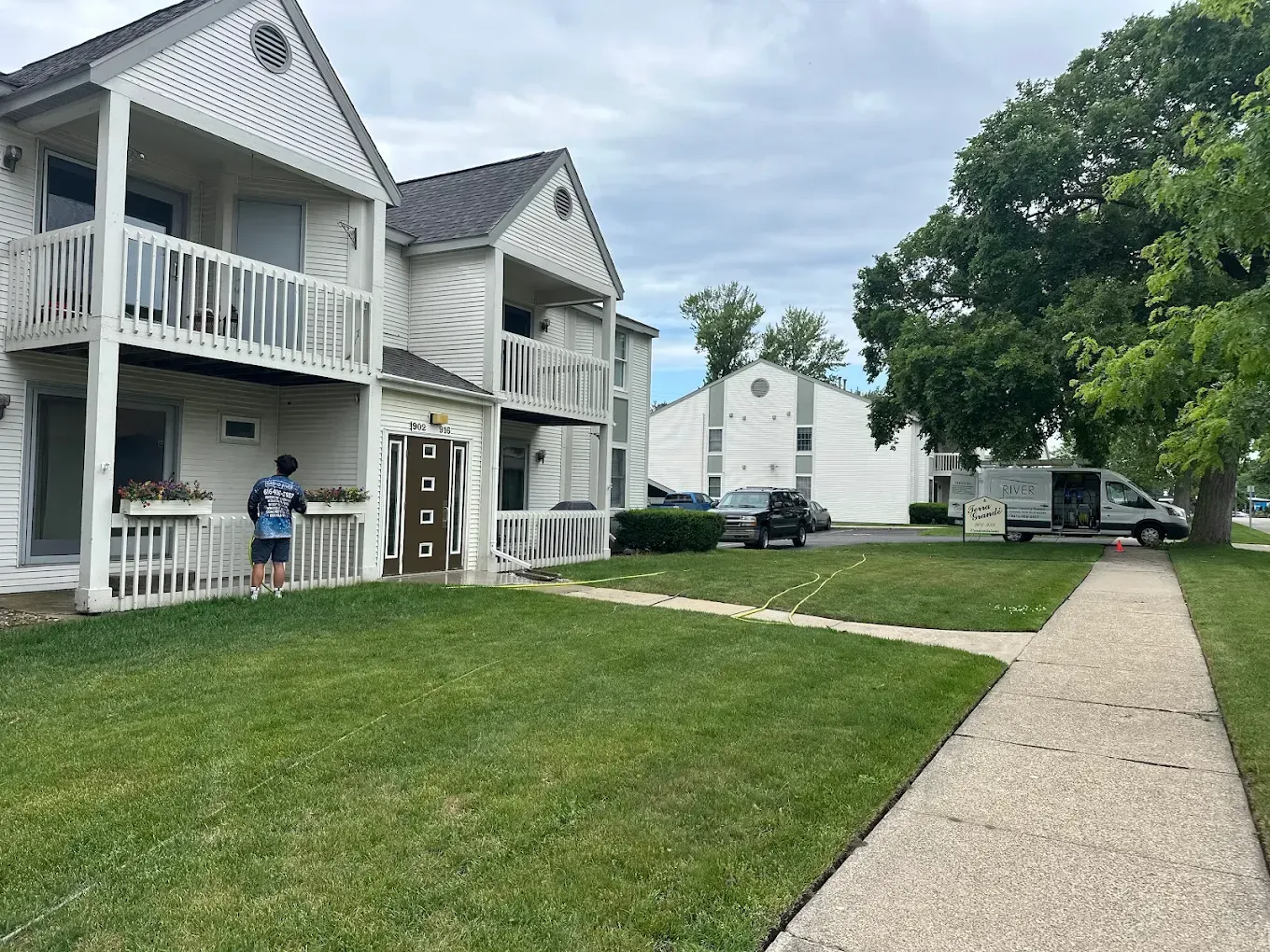 Two-story white apartment buildings with balconies, a grassy lawn, and a person loading a van.