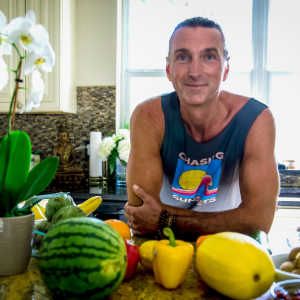 Man smiling behind a kitchen counter with fresh fruit and vegetables, including watermelon, mango, and orchids in a pot