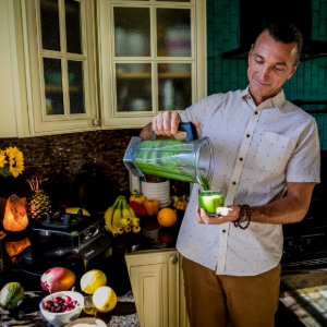 Man in a kitchen pouring green smoothie into a glass beside fruit, flowers, and a blender