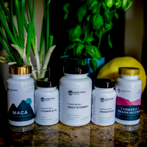 Five white supplement bottles on a countertop with green plants and a yellow bowl in the background