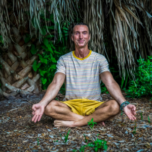 Person meditating cross-legged on the ground before a thatched wall, wearing a striped shirt and yellow shorts.