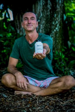 Person sitting cross-legged outdoors, holding out a small white bottle toward the camera.