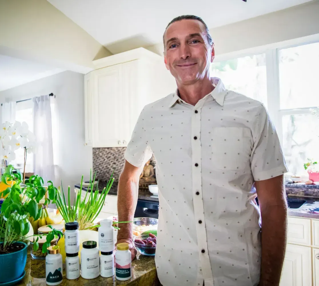 Man standing in a bright kitchen beside potted plants and bottles on the counter