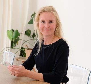 Een vrouw zit aan een tafel met een helder glas in haar hand, met een potplant en witte gordijnen achter haar.