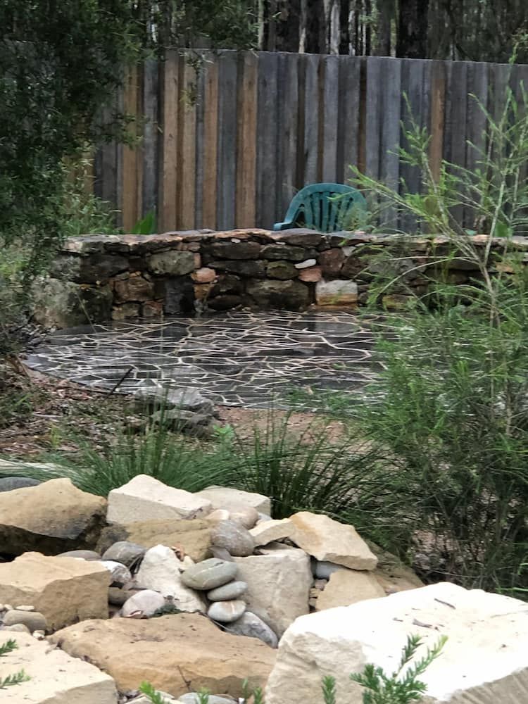 A Rock Wall With a Blue Chair in the Background and a Wooden Fence — Peter Phillips Landscape Design in Bomaderry, NSW