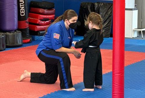 A patient and kind instructor at American Tiger Martial Arts in Westminster, CA is kneeling down next to a shy little girl in a karate uniform. They are working on her confidence together.