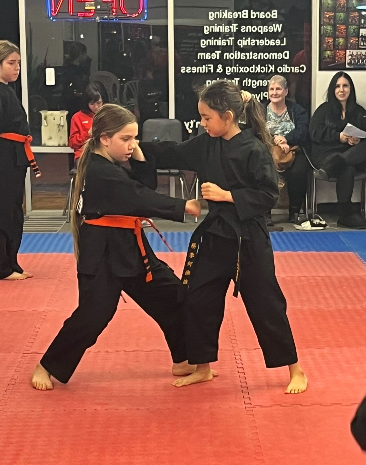 Two martial arts students spar on red mats in a dojo, wearing black uniforms with orange belts.