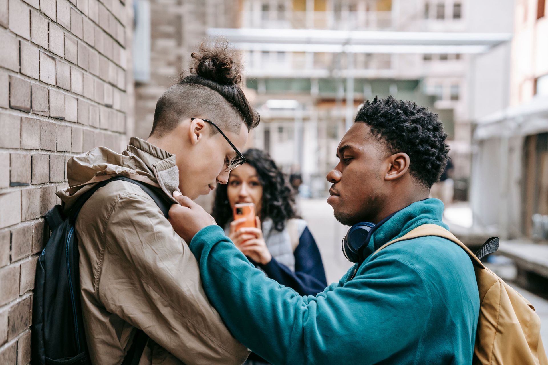 One person comforts another against a brick wall while a third stands nearby holding a phone.