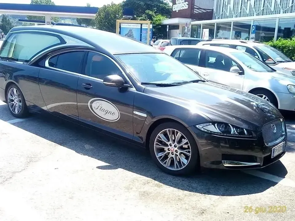 Carro funebre Jaguar grigio scuro parcheggiato, con logo ovale sul lato, vicino ad altre auto e a un edificio.