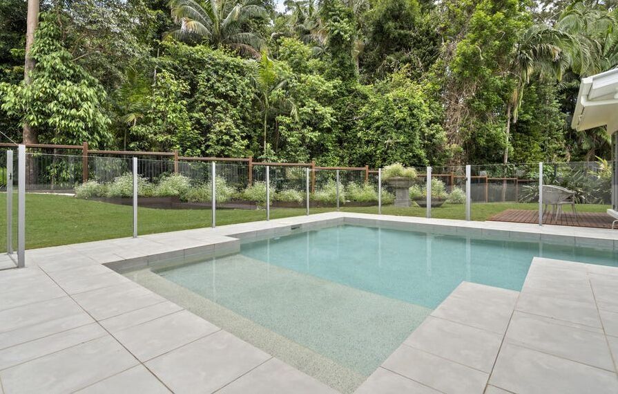 Backyard Pool With Glass Safety Fence and Light Stone Deck — Mad Pool Interiors and Renovations in Baringa, QLD