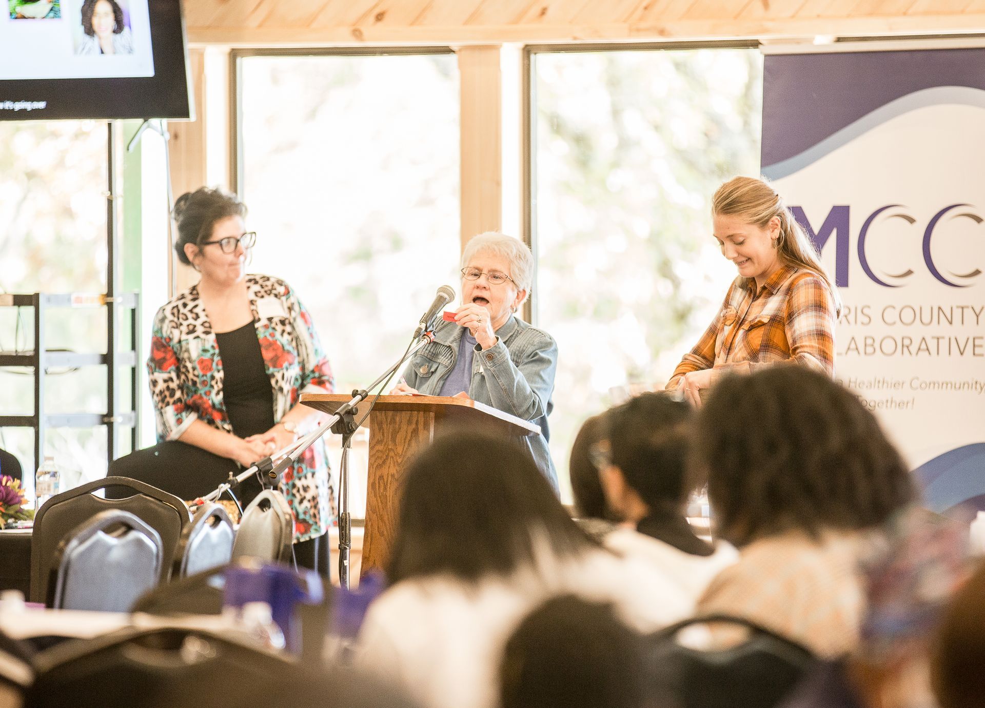 Two women are sitting at a podium talking to a crowd of people.