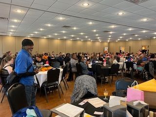 A man is standing in front of a large room filled with people sitting at tables.