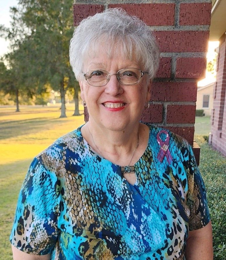 A woman wearing glasses and a blue shirt is standing in front of a brick wall.