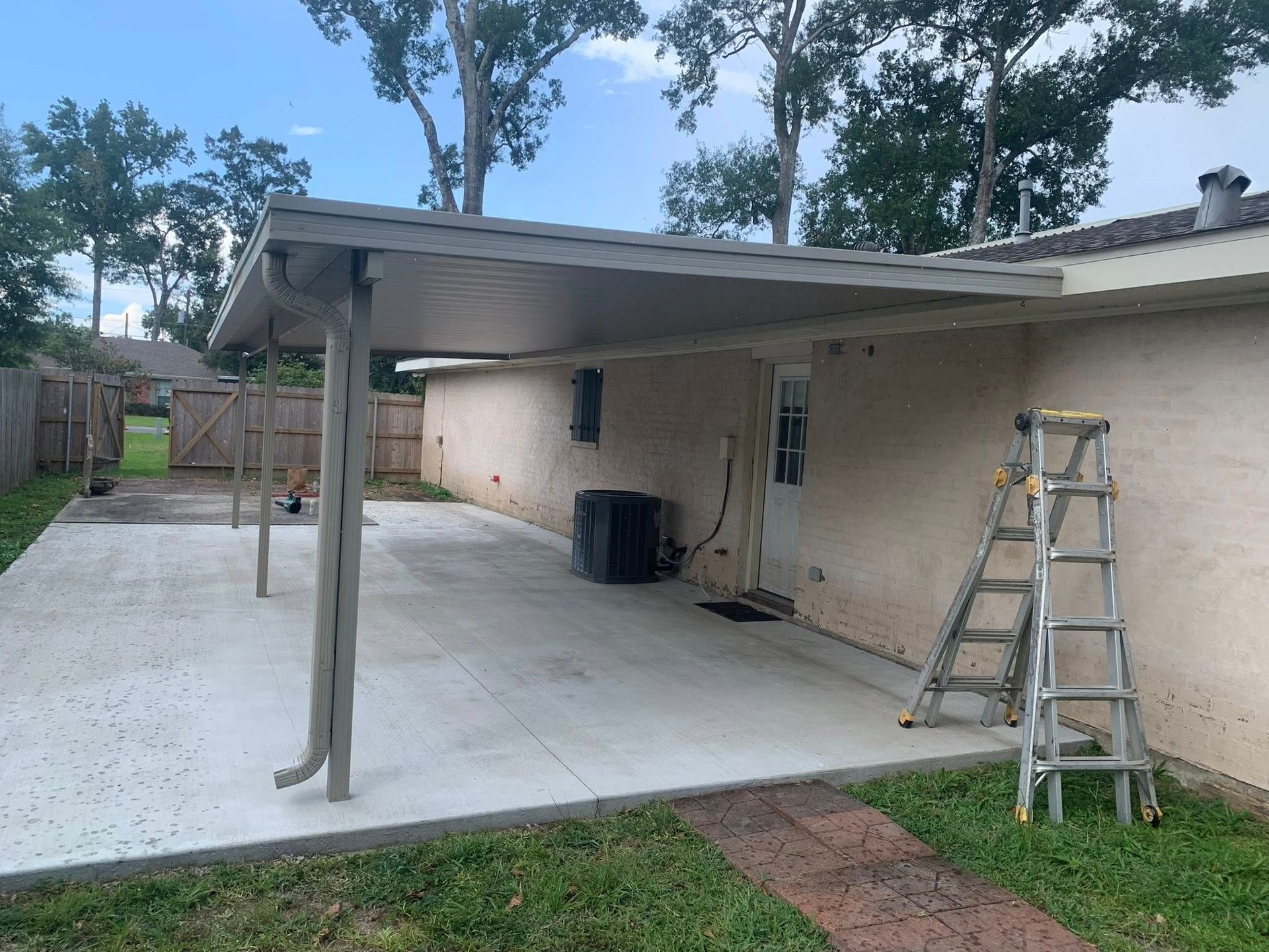 Backyard patio with a covered carport, ladder, fence, and concrete slab beside a beige house.