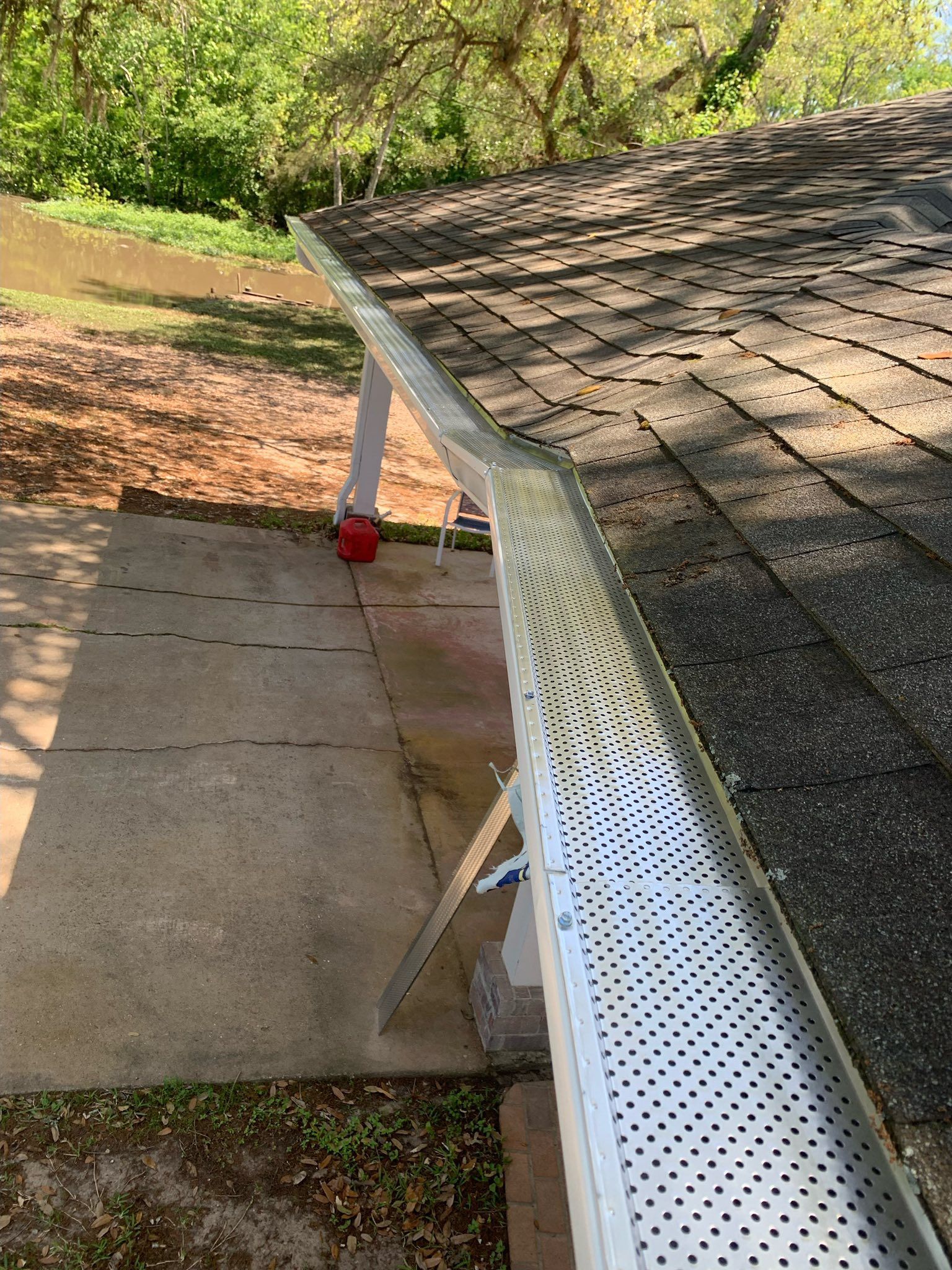 Roof with white gutter and downspout beside a concrete patio, with trees and grass in the background