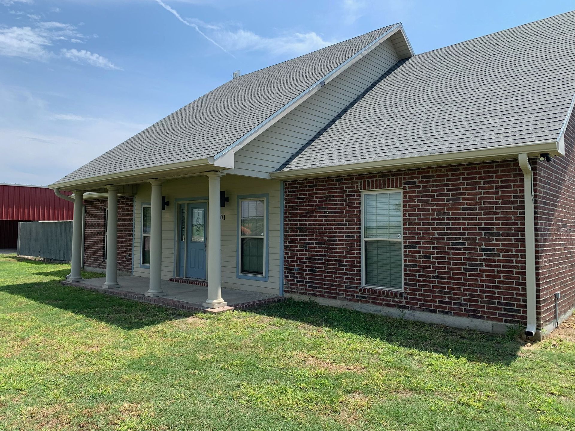 Single-story brick house with gray roof, white columns, and a green lawn under a clear sky