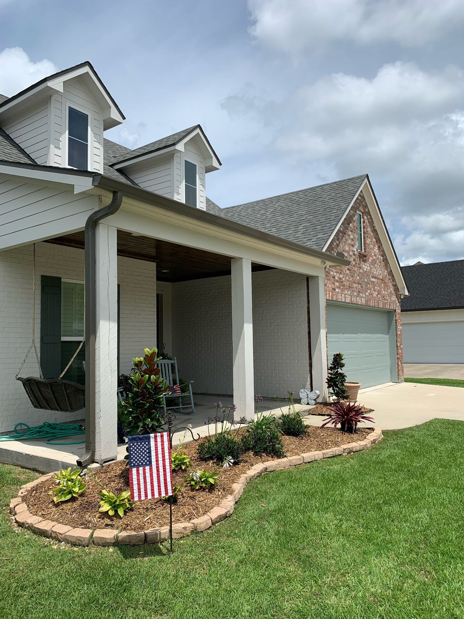 Front porch of a suburban house with white columns, flower bed, and small American flag on the lawn.