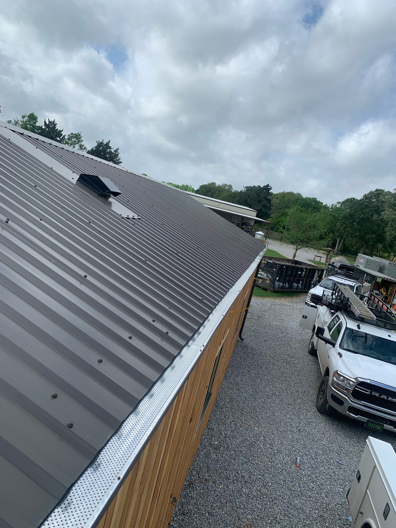 Gray metal roof with a skylight beside a gravel driveway and parked cars under cloudy skies