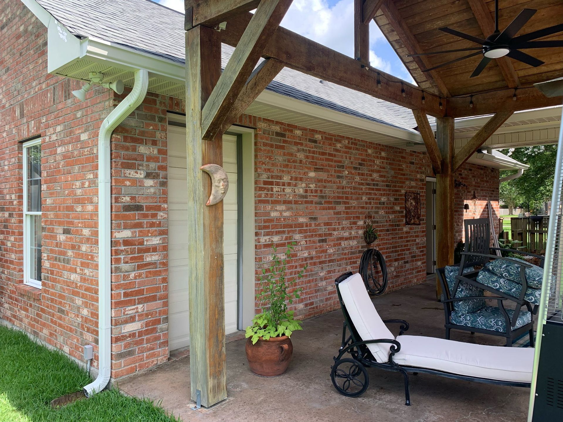 Covered brick patio with wicker furniture, lounge chair, and potted plant under a wooden pergola