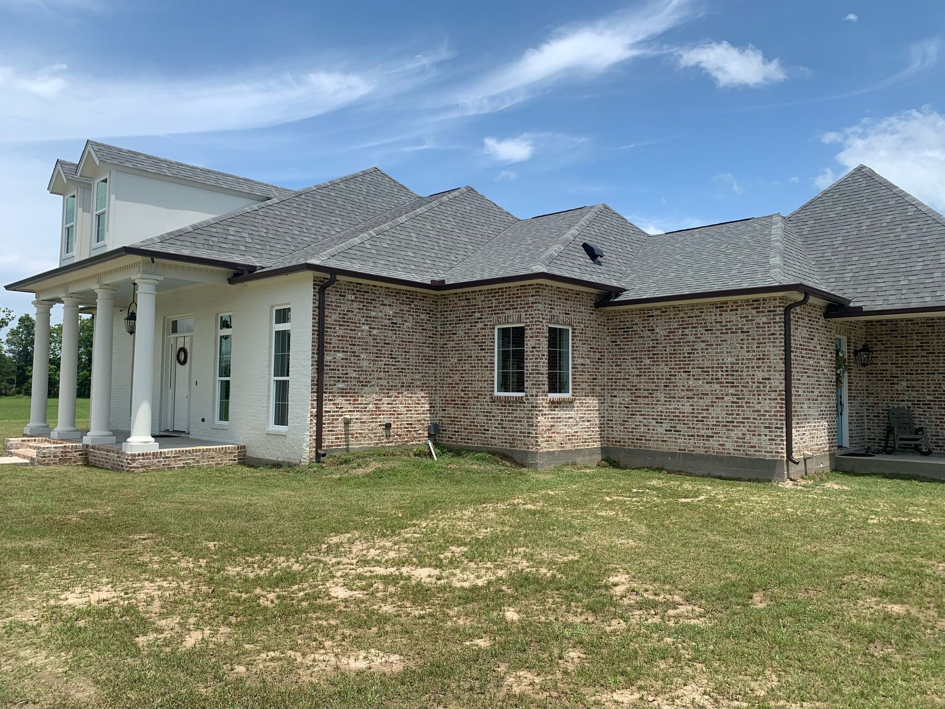 Single-story brick house with white columns and a gray roof on a grassy lot under a blue sky