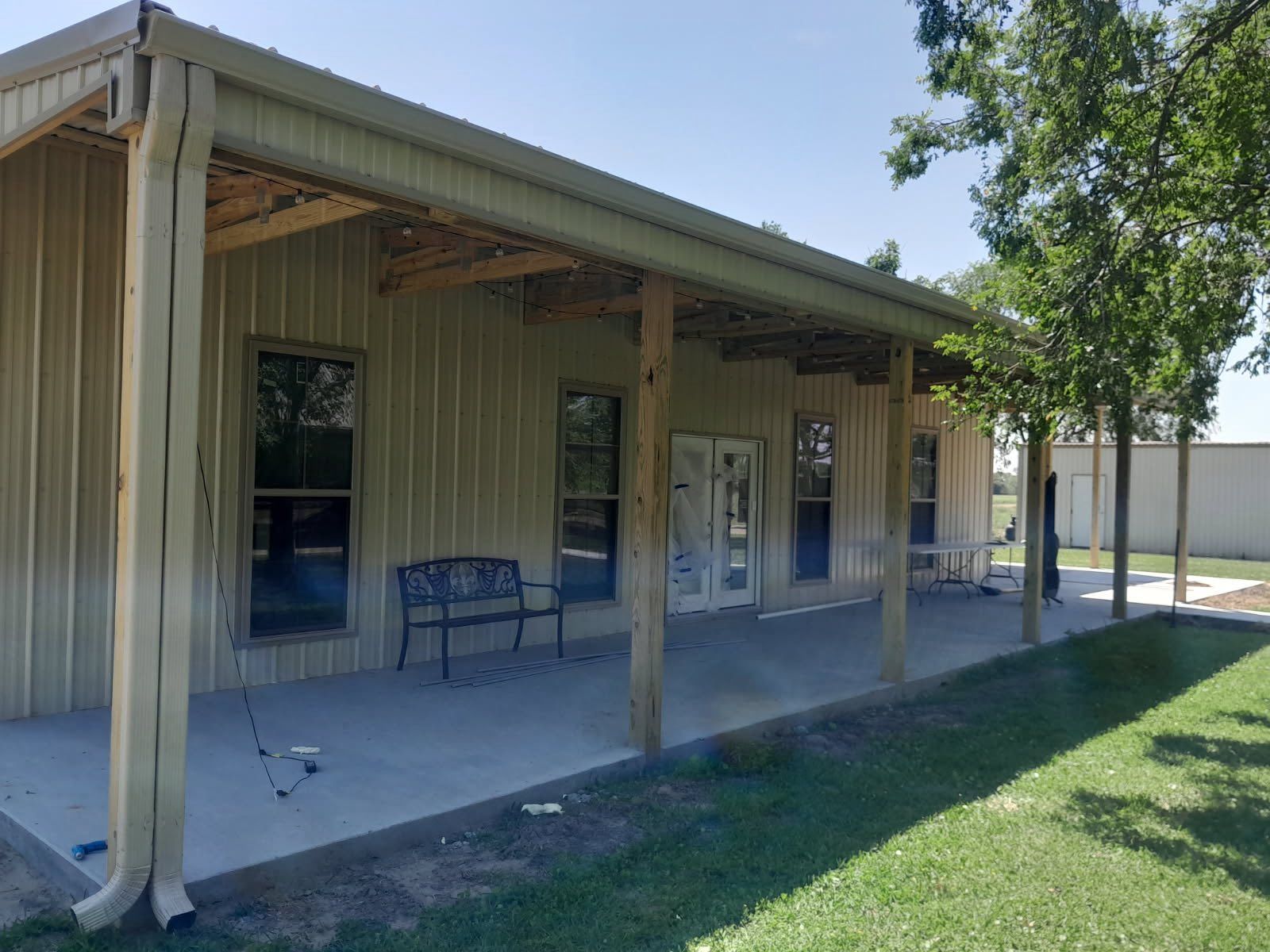 Covered porch on a beige building with benches, windows, and a grassy yard beside a tree.