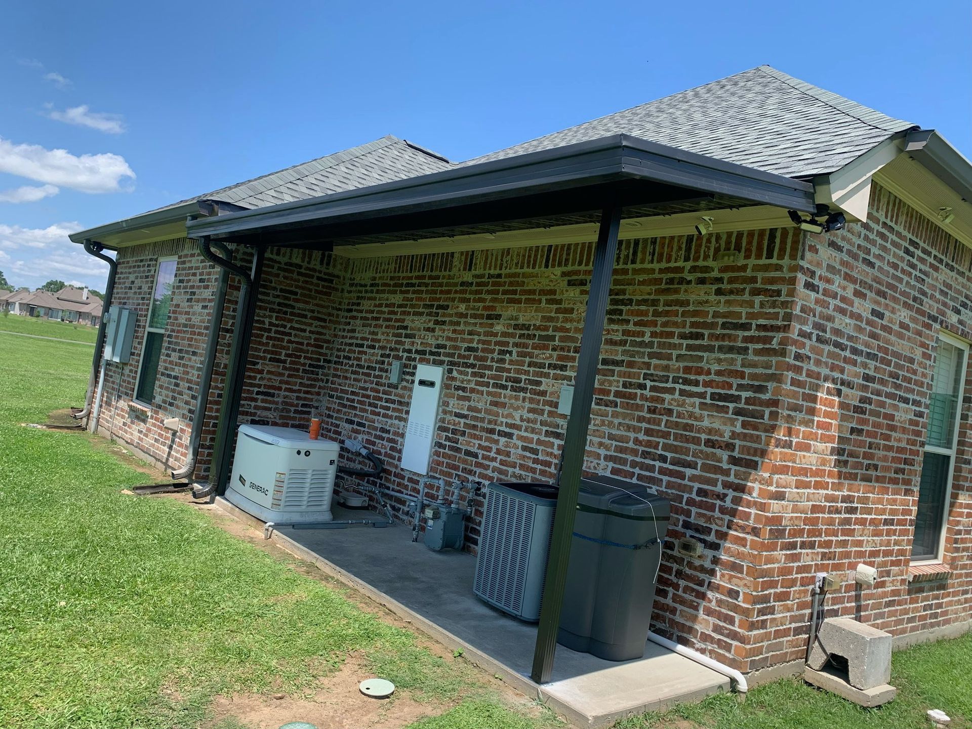 Brick house exterior with covered patio, HVAC unit, trash bins, and lawn under a blue sky