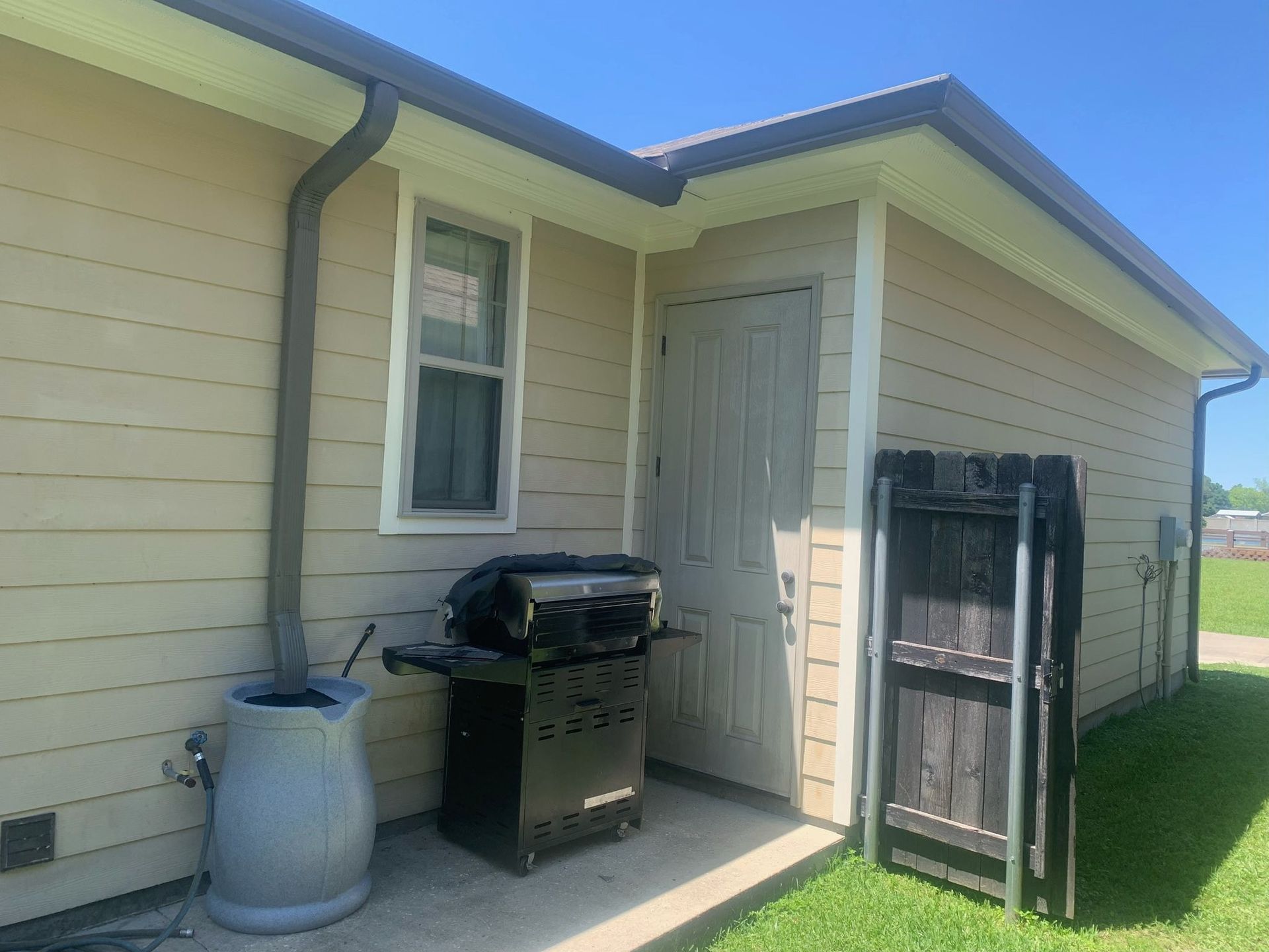 Side yard patio with a grill, trash can, and folded gate beside a beige house under a clear sky