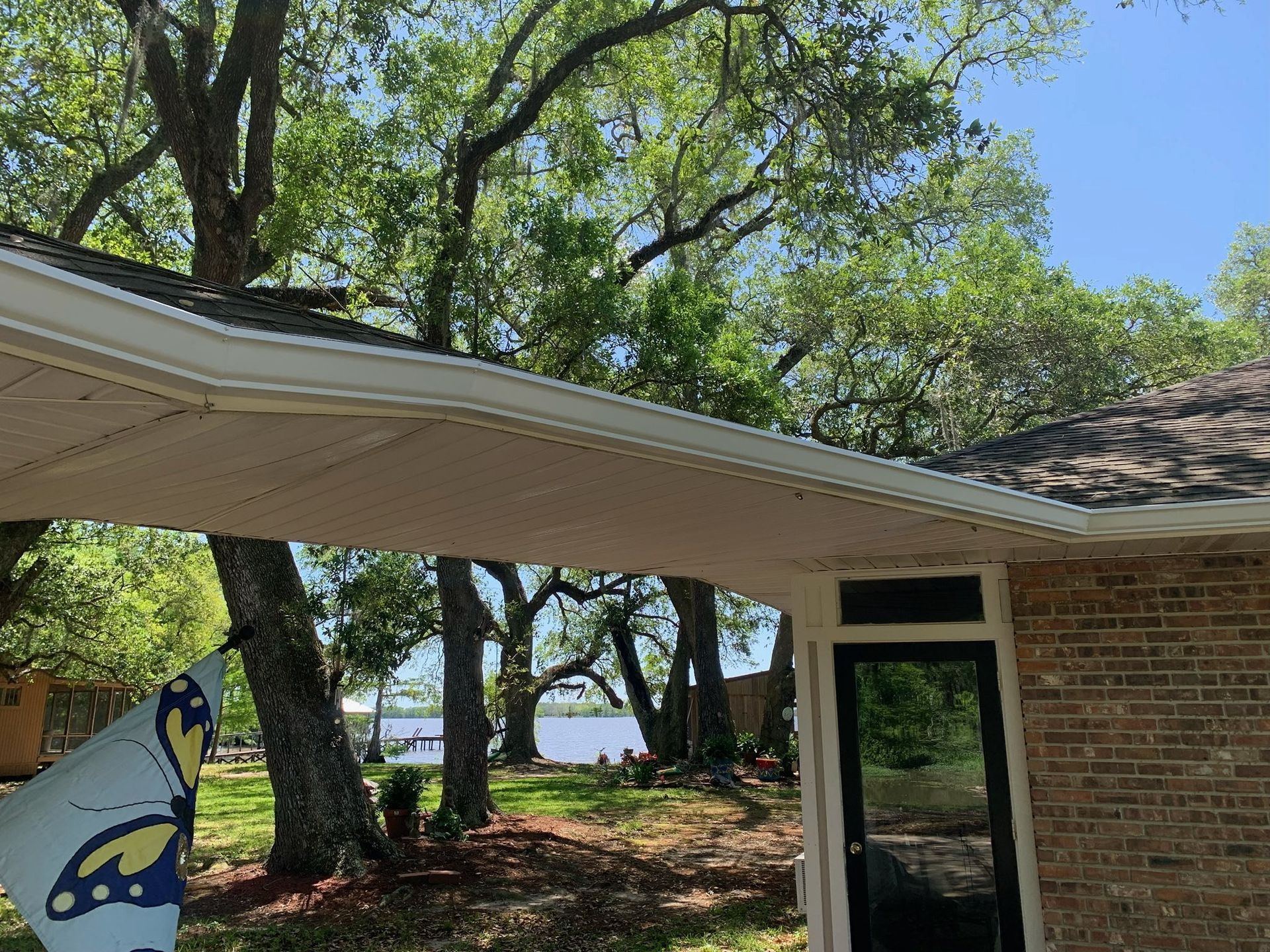 Brick house patio with screened door, covered roof, and large trees by a waterfront lawn