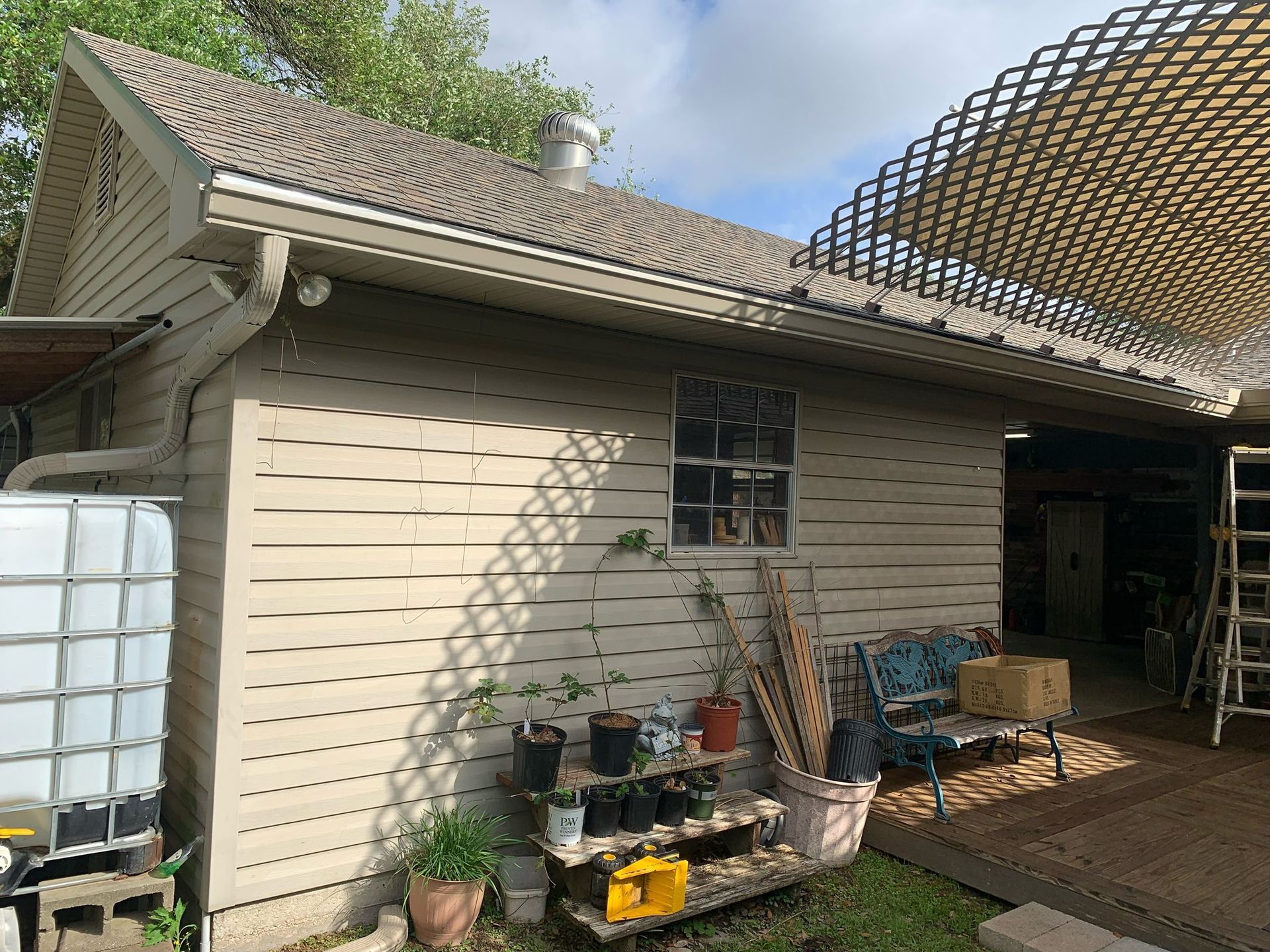 Backyard shed with beige siding, potted plants, stacked ladders, and a wooden deck under a lattice roof