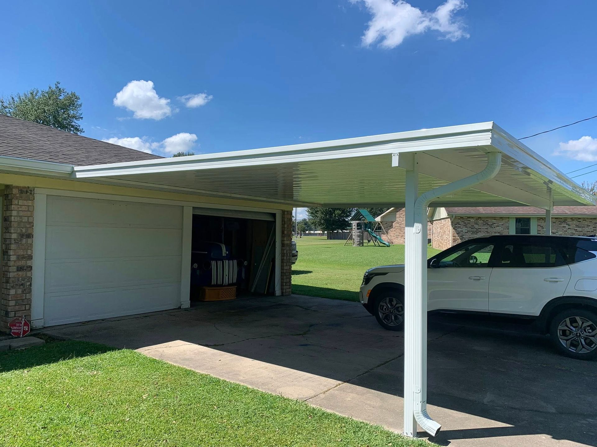 White car under a metal carport beside a house with a green lawn and blue sky