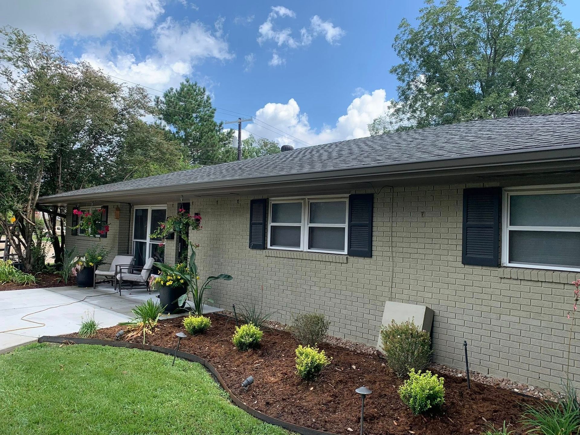 Single-story beige brick house with black shutters, a front porch, and a landscaped lawn under a blue sky
