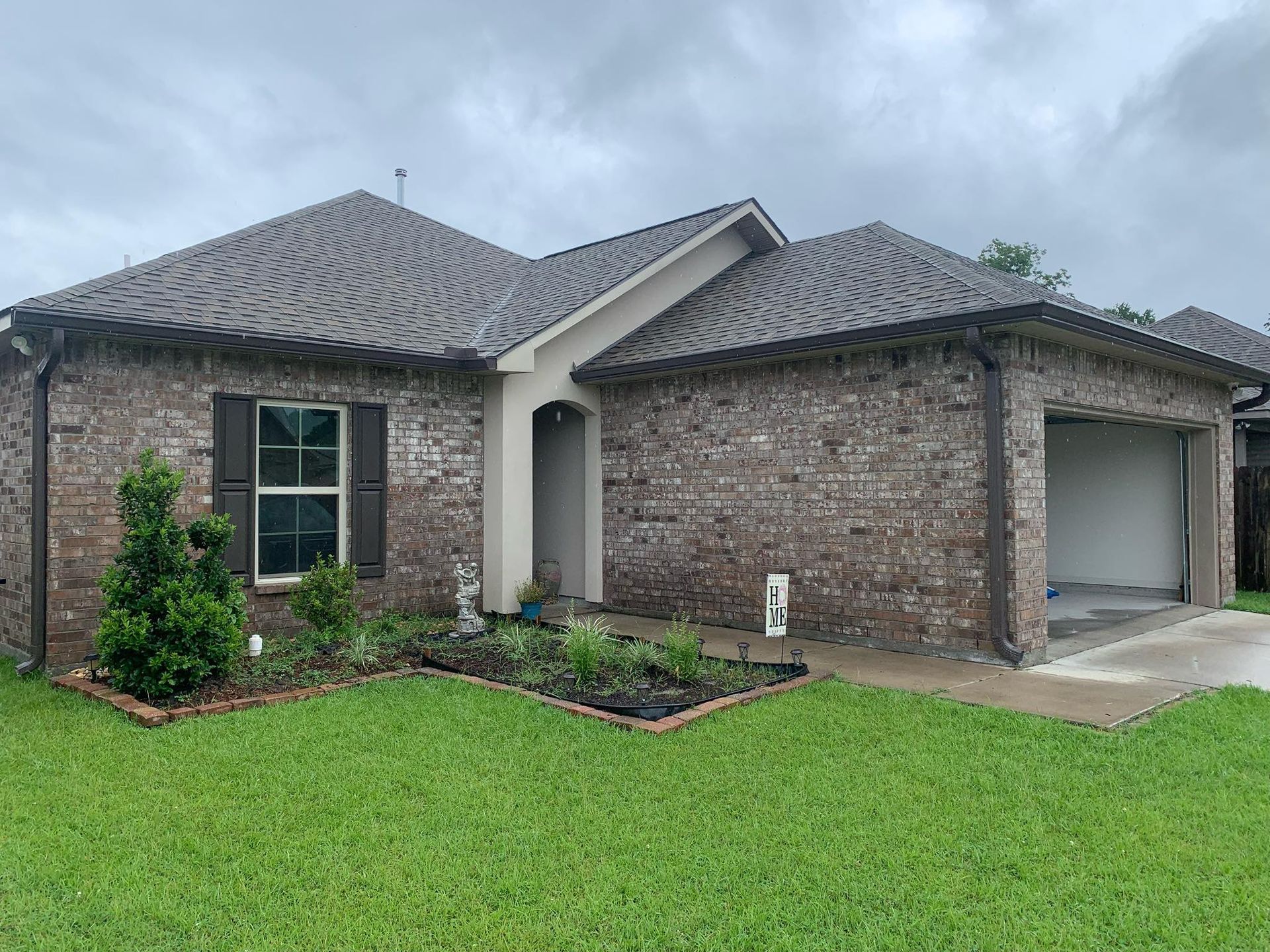 Single-story brick house with gray roof, green lawn, and attached garage under an overcast sky