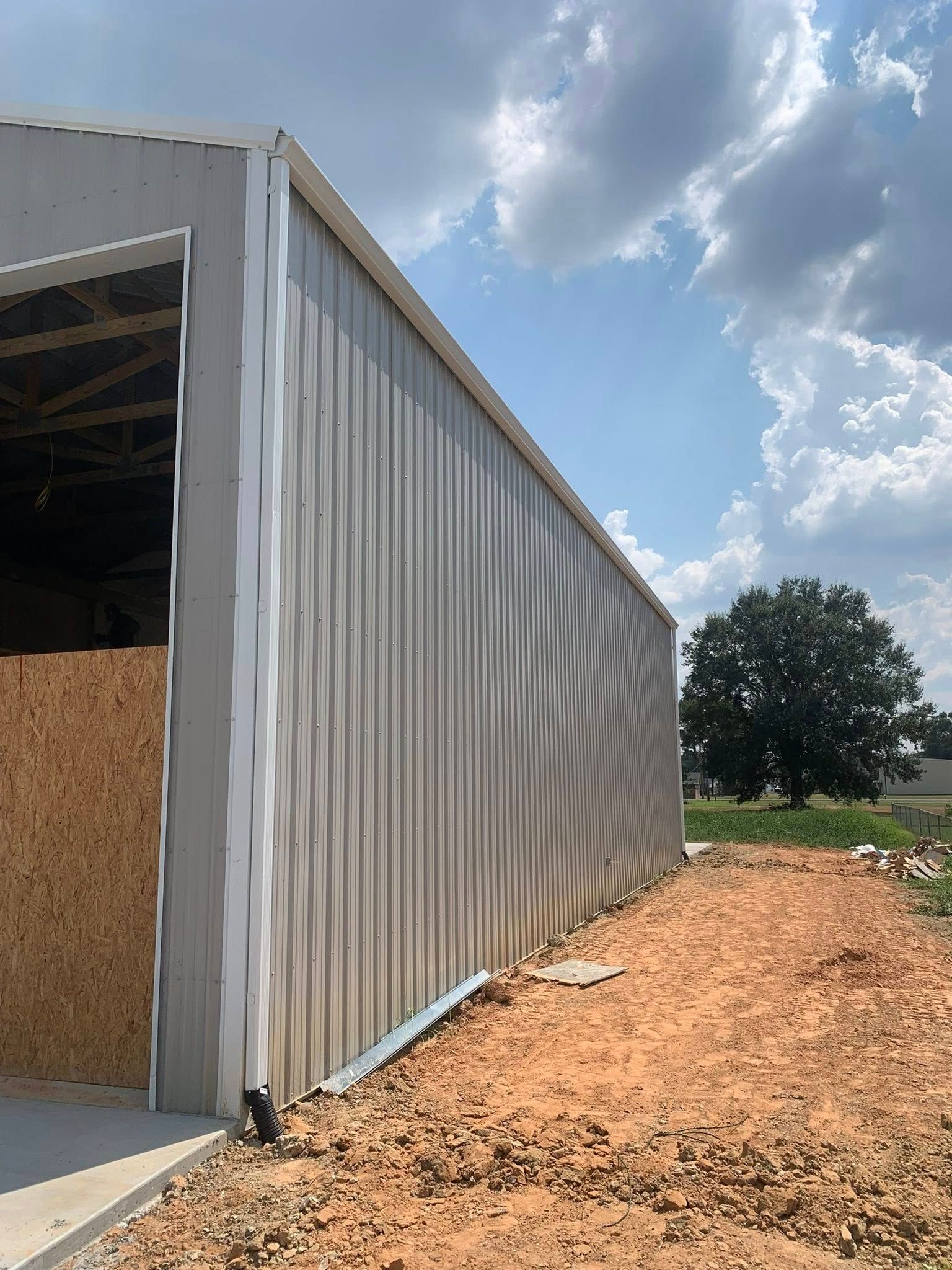 Metal-sided barn or shed under construction on a dirt lot beside a tree under a cloudy sky