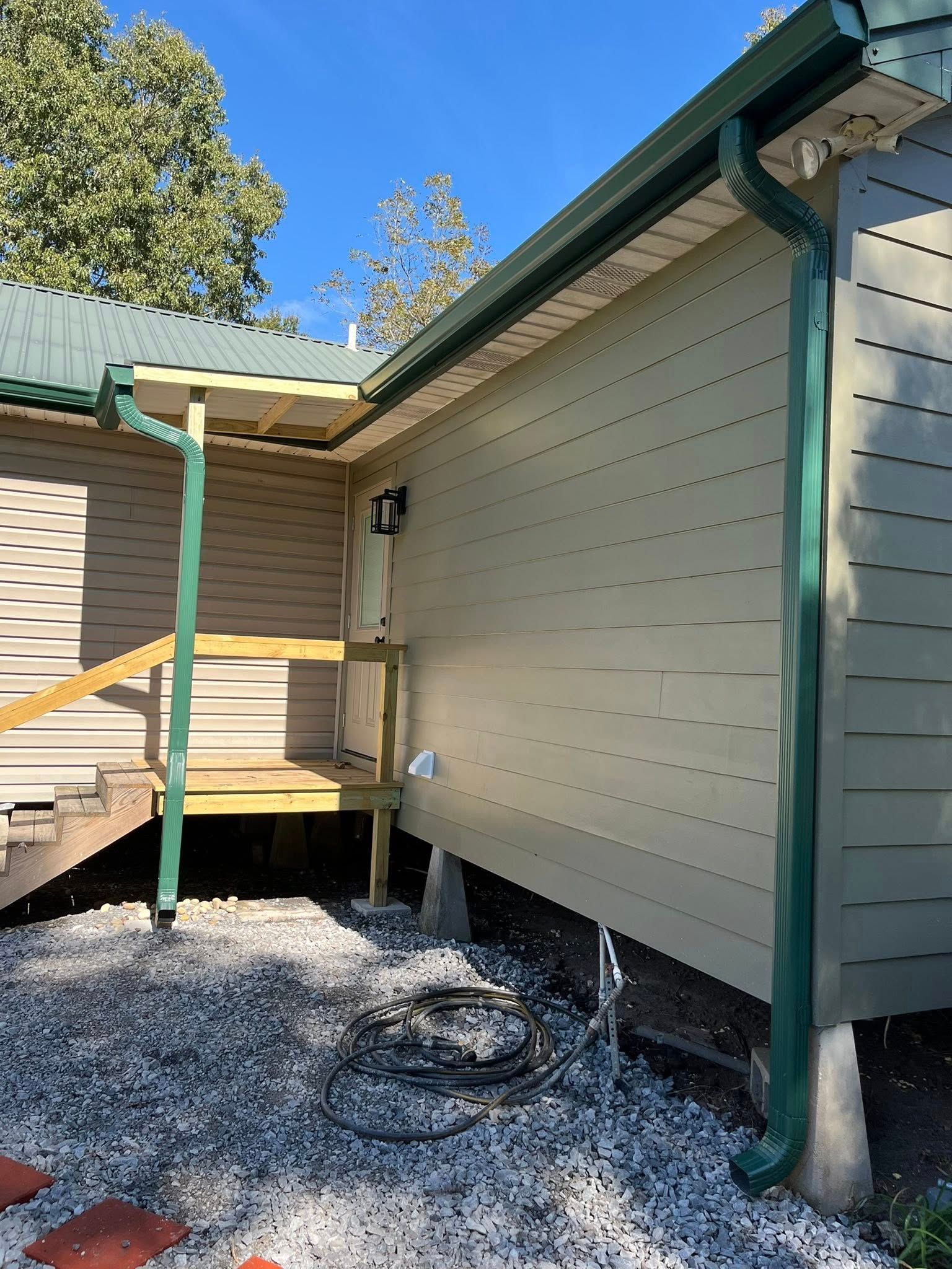 Exterior side of a beige house with green trim, raised deck, downspout, and gravel ground