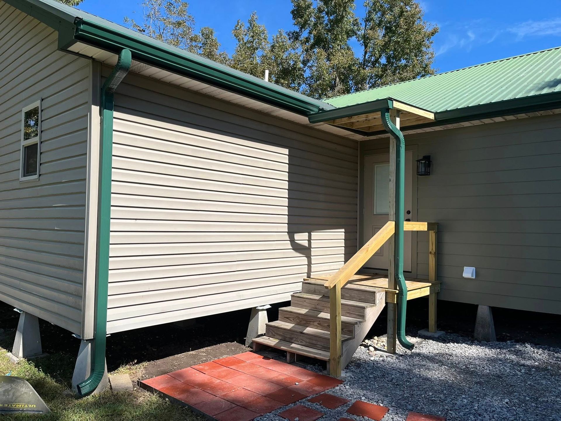 Small house exterior with green roof, beige siding, and steps leading to a side entrance.