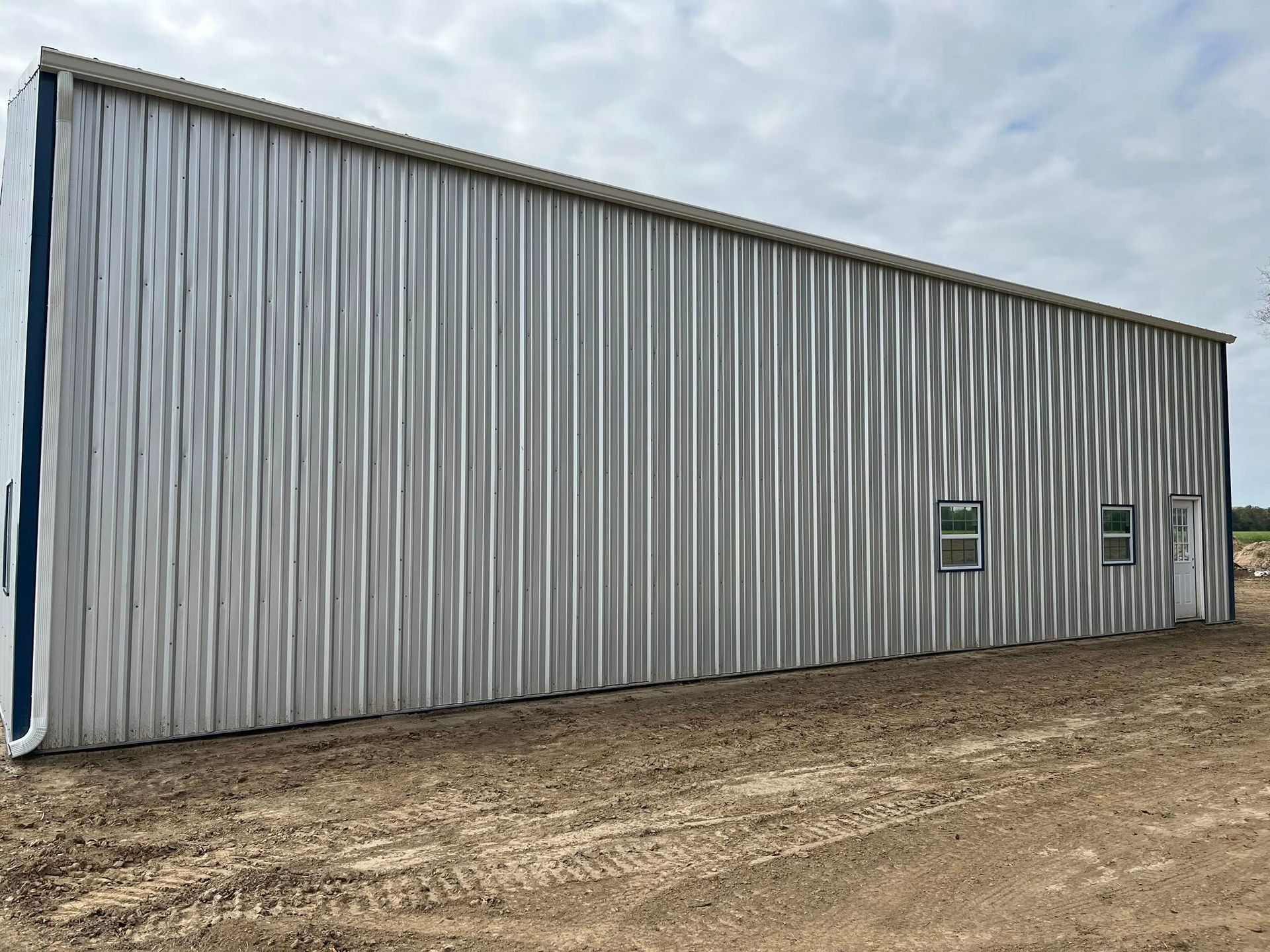 Large gray corrugated metal warehouse with two small windows on a dirt lot under a cloudy sky