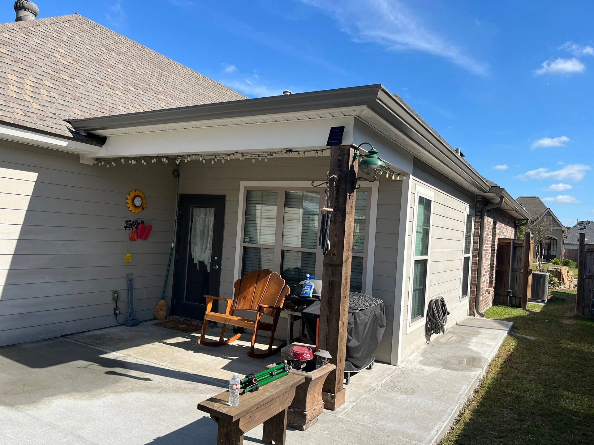 Back patio of a house with wooden chairs, potted plants, and a covered walkway under a blue sky