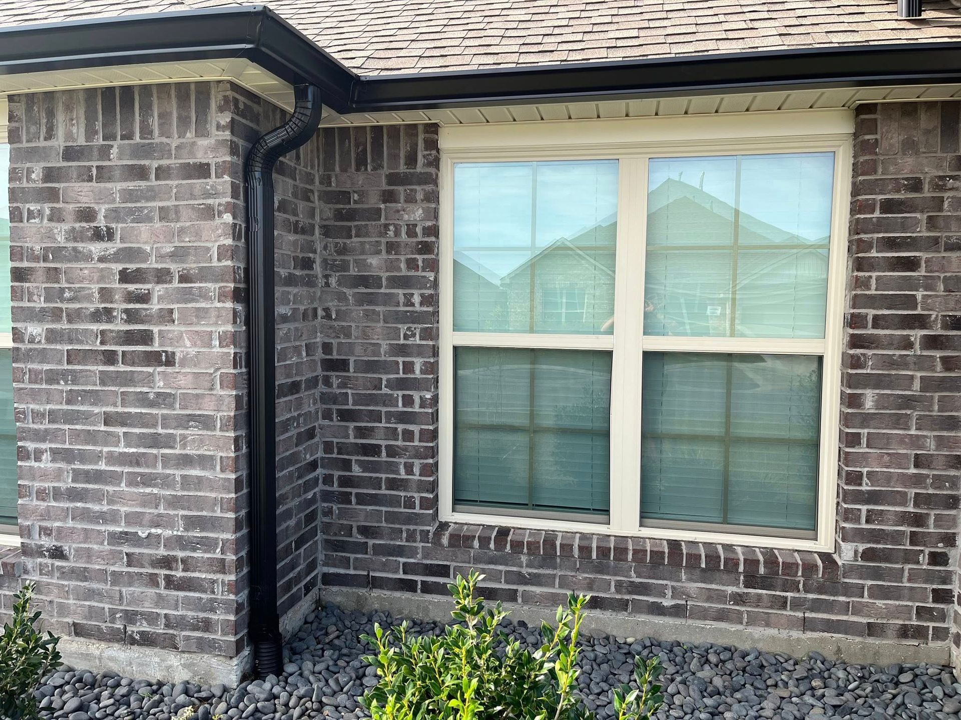 Brick house exterior with a white-framed window and black downspout beside a small shrub bed