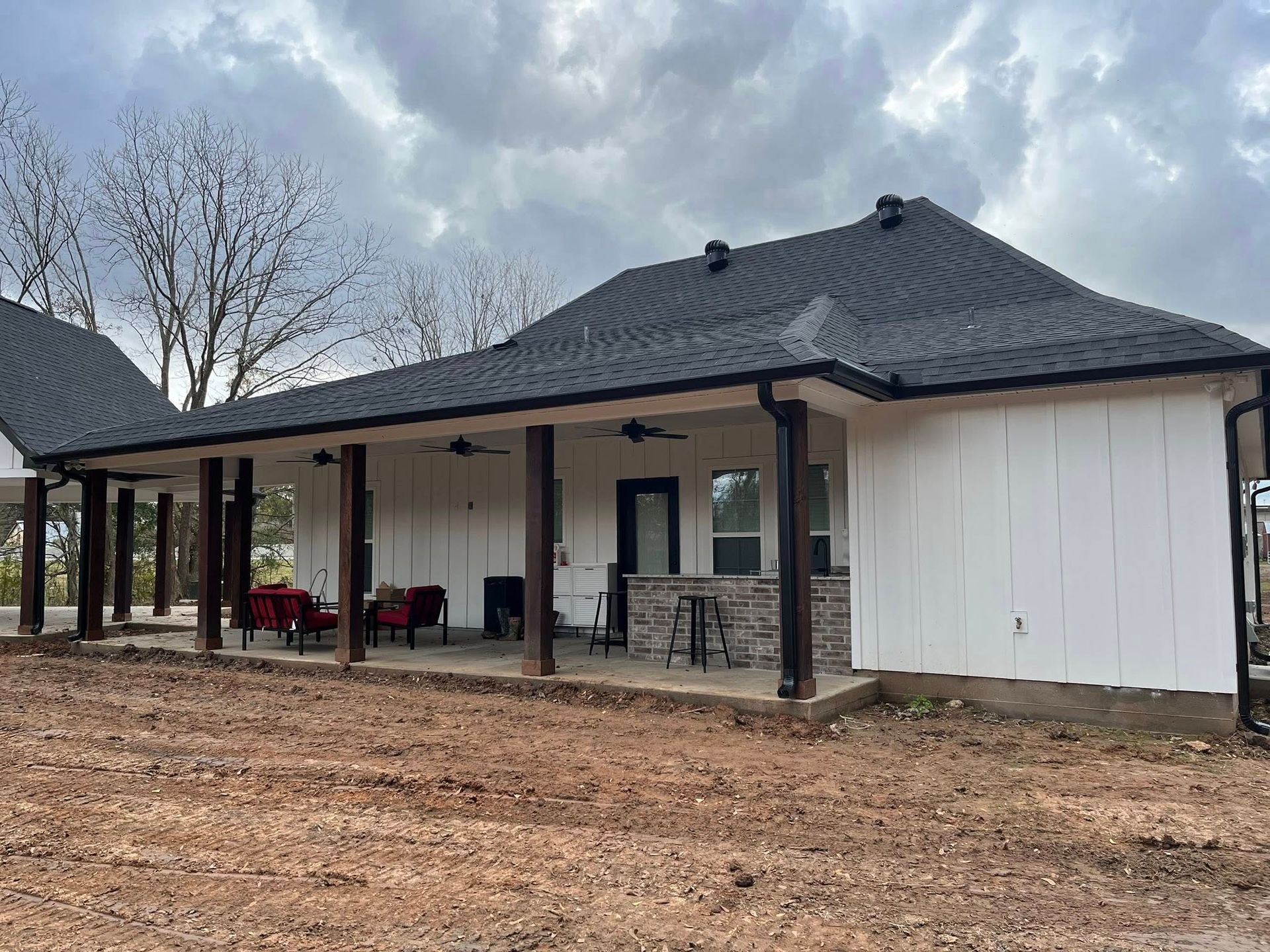 Modern white house with black roof and covered porch on a dirt lot under cloudy sky