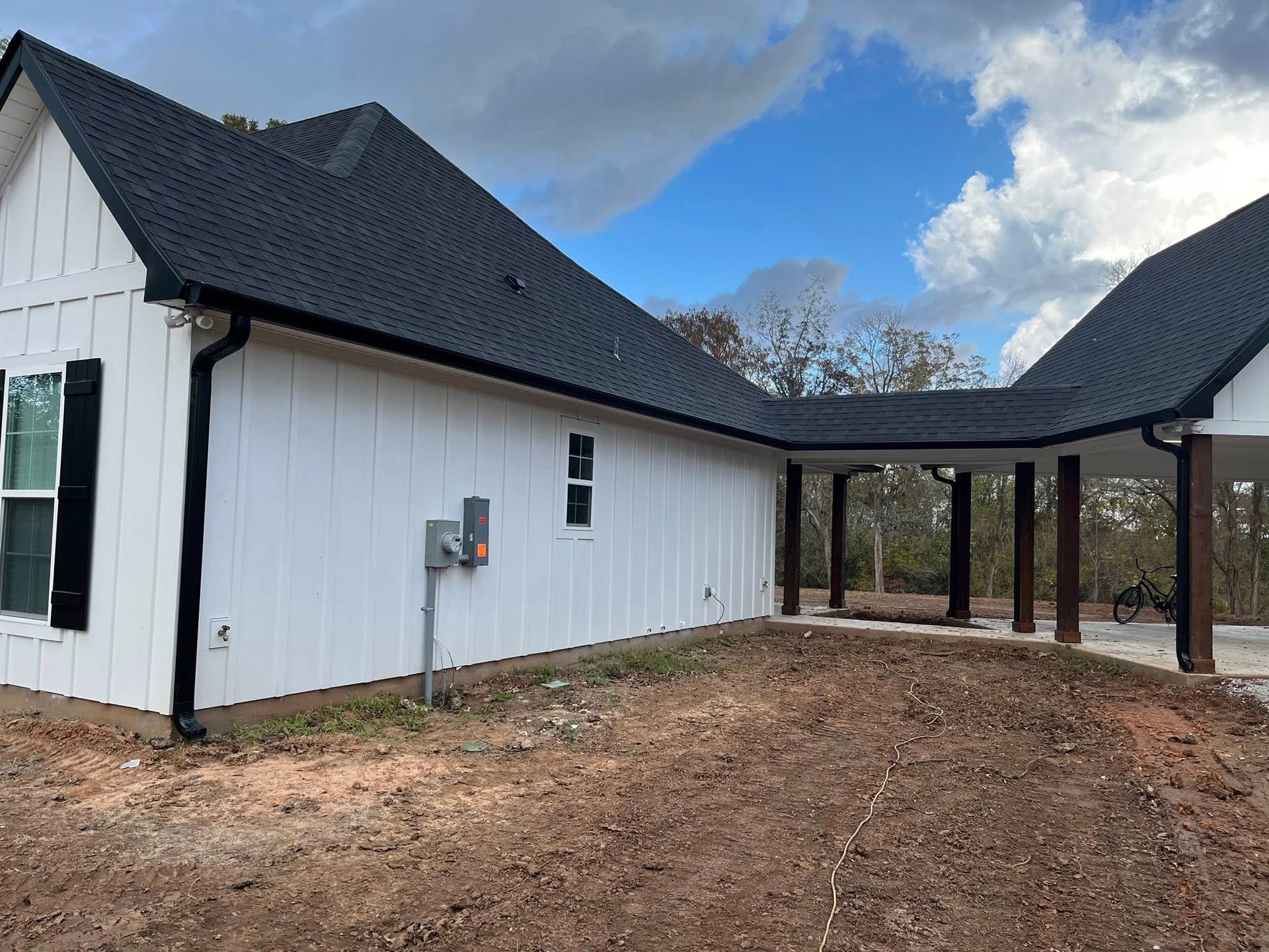 White house exterior with black roof and covered carport on a dirt lot under a cloudy blue sky